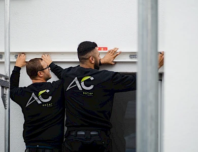Zwei Mitarbeiter von AC Solar installieren technische Komponenten einer Photovoltaikanlage.